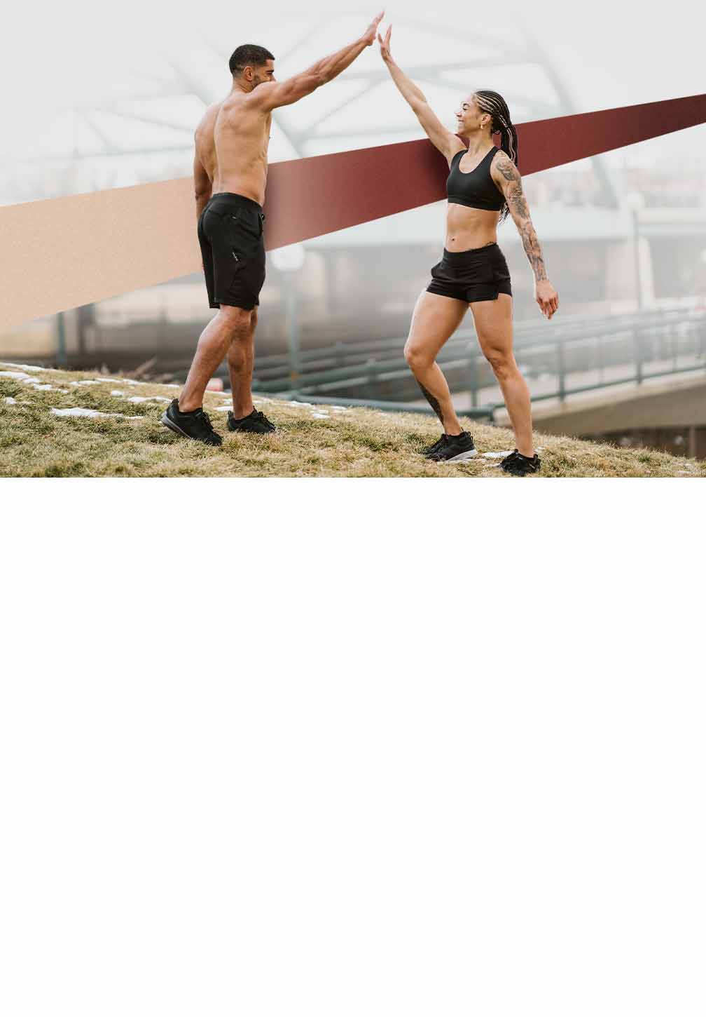 A muscular, shirtless man and a woman with an arm tattoo, both wearing Hylete black shorts, enthusiastically high-five outdoors on a grassy hill in a cool-weather setting.